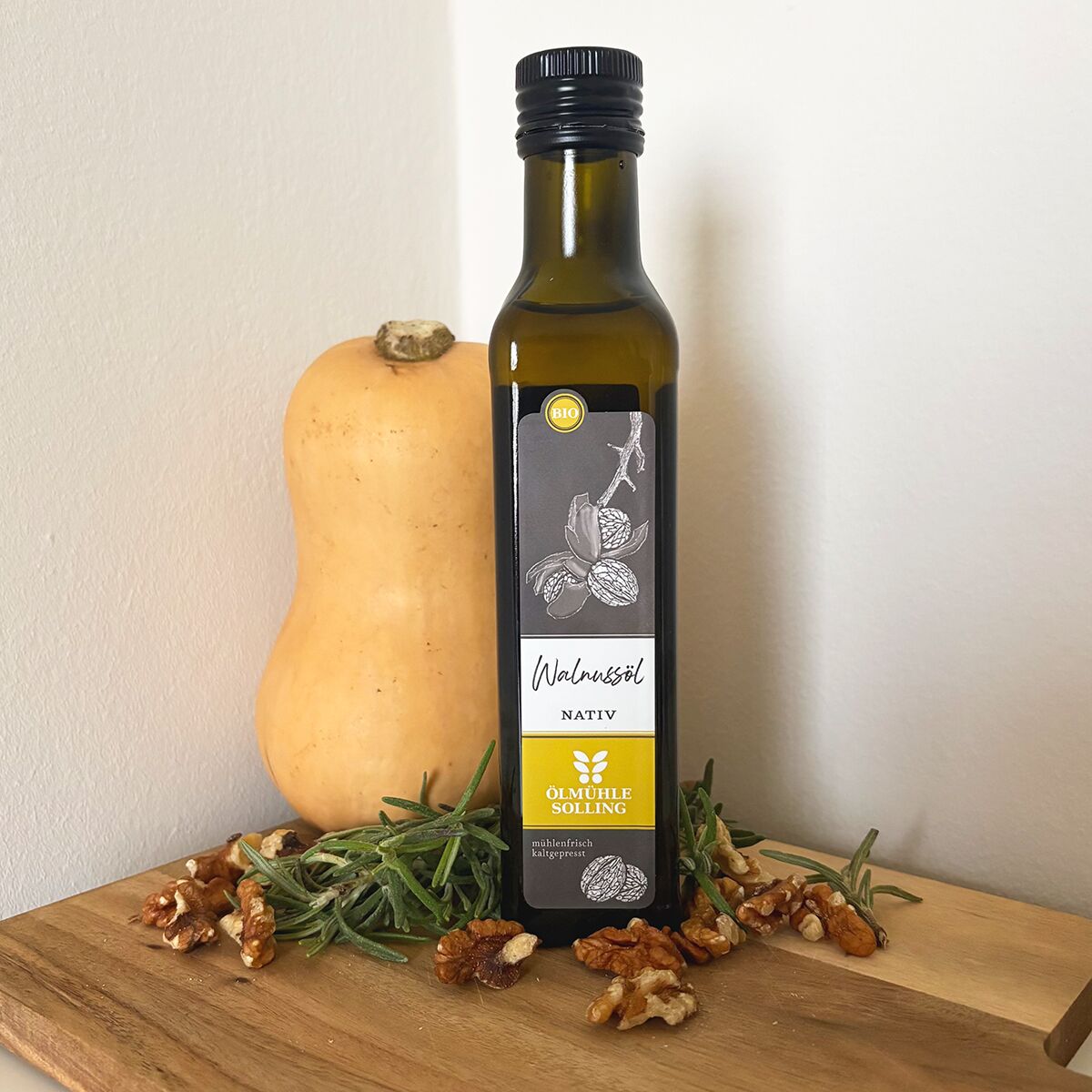 Eine Flasche natives Bio-Walnussöl der Marke Ölmühle Solling steht auf einem Holzbrett. Daneben liegen Walnüsse, frische Kräuter und ein kleiner Butternut-Kürbis vor einer hellen Wand. Das Arrangement wirkt natürlich und einladend, ideal für Genießer gesunder, hochwertiger Lebensmittel.