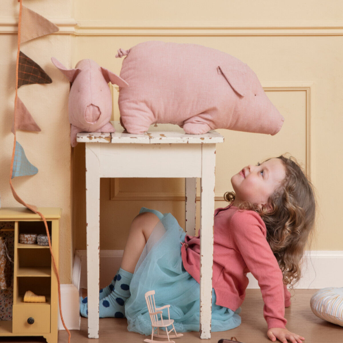 Ein großes Kuscheltier in Form eines rosa Schweins sitzt auf einem weißen Tisch in einem Kinderzimmer. Ein kleines Mädchen mit lockigem Haar und einem rosa Pullover schaut zu dem Schwein hoch und hält ein Spielzeugboot in der Hand. Der Hintergrund des Zimmers ist in warmen Beige- und Pastellfarben gehalten.
