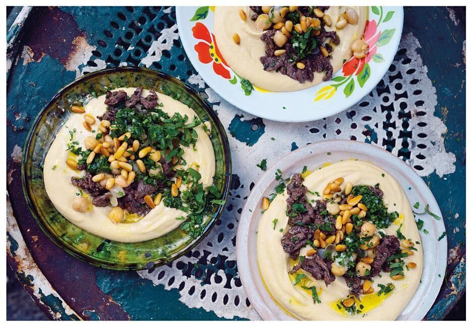 Drei Teller mit cremigem Hummus, garniert mit Pinienkernen, Kräutern und Gewürzen auf einem dekorativen Tisch.