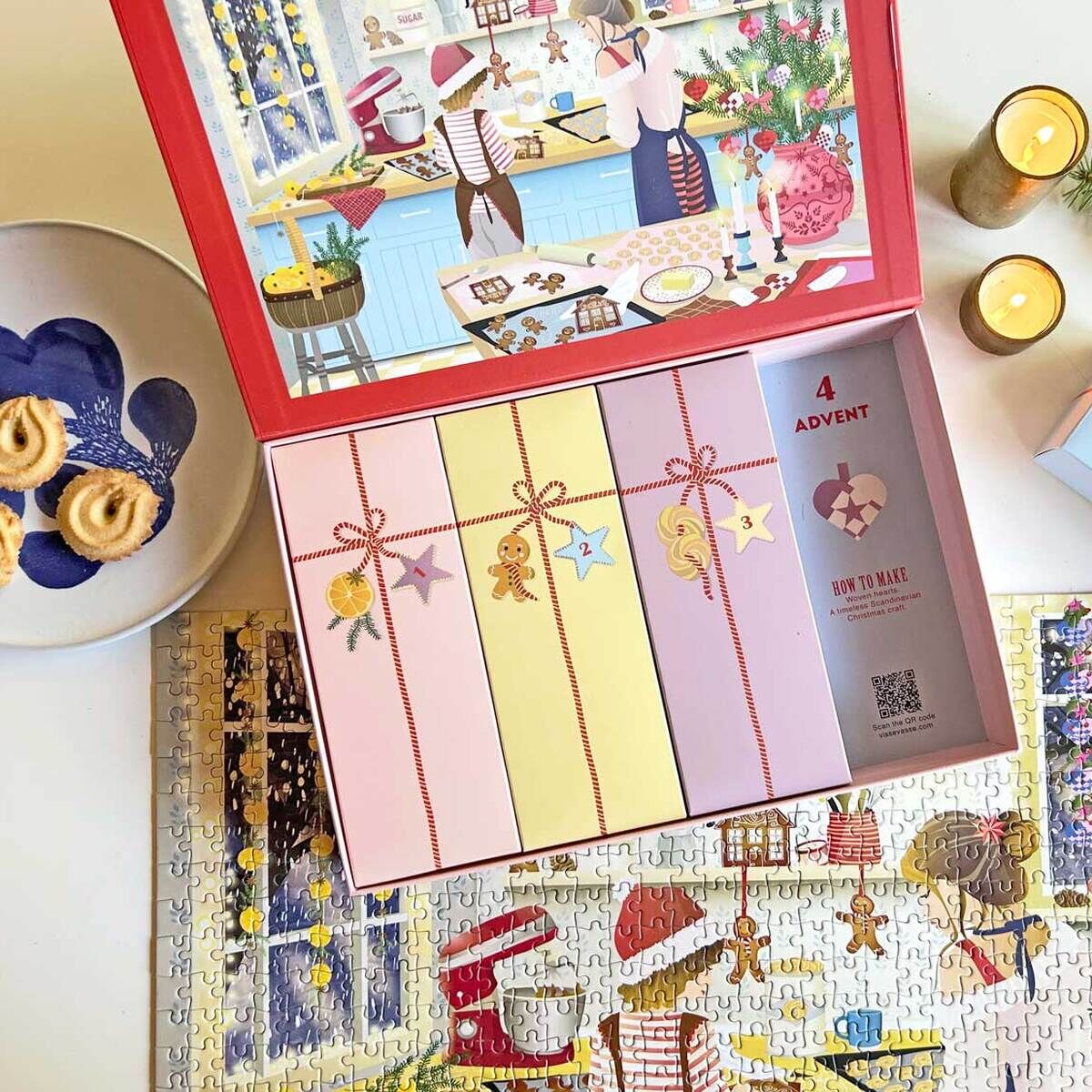 Puzzle-Adventskalender »Sweet Christmas« von VISSEVASSE, geöffnete Schachtel mit bunten, nummerierten Fächern, liebevoll illustriertem Küchenmotiv, aufgebautem Puzzle mit Weihnachtsbild, daneben Keksteller, Weihnachtszweig, Kerzen und Geschenkbox.