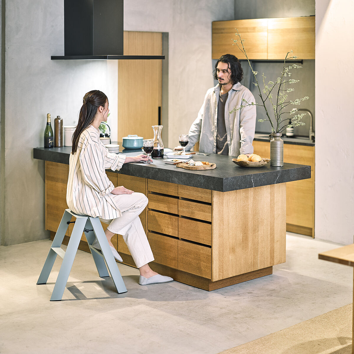 Eine moderne, zweistufige Lucano Trittleiter steht in einer hellen, minimalistischen Küche neben einer Kücheninsel. Eine Frau sitzt entspannt auf der Trittleiter und unterhält sich mit einem Mann, der an der Kücheninsel steht. Die Küche ist mit hellen Holzmöbeln und neutralen Farbtönen gestaltet.