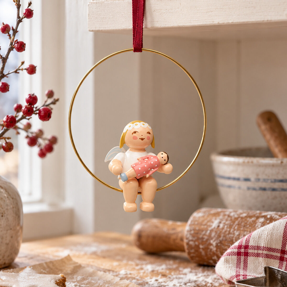Ein hölzerner Christbaumengel von Wendt & Kühn sitzt in einem goldenen Ring und hält eine Puppenfigur. Im Hintergrund sind Backutensilien, Sterne aus Gebäck, ein Tuch, ein Steingut-Topf und weihnachtliche Deko auf einer Holzoberfläche zu sehen.