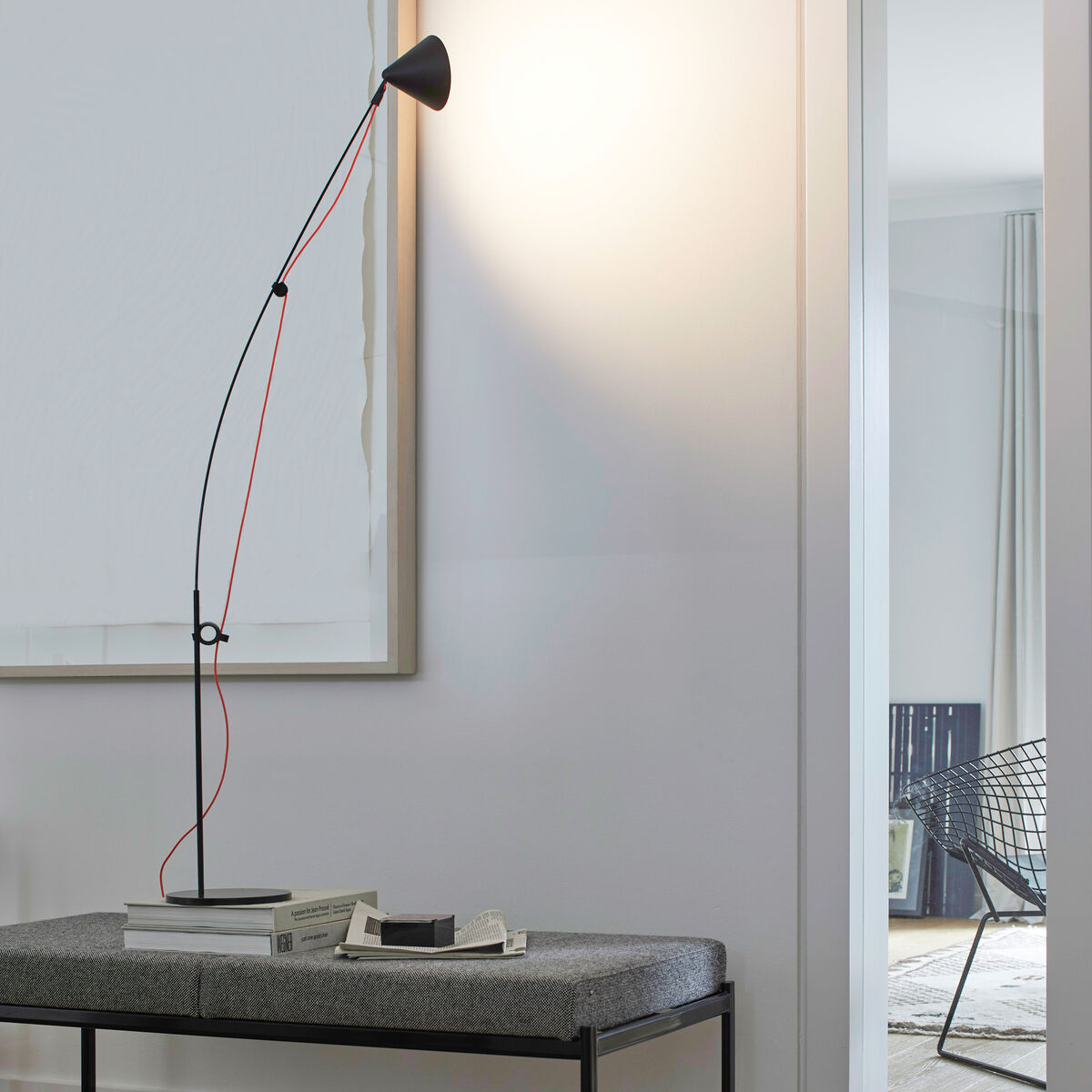 Eine moderne Tischlampe »AYNO« mit einem schlanken, verstellbaren schwarzen Arm und einem konischen Lampenschirm. Die Lampe steht auf einem minimalistischen Tisch in einem hellen Raum. Ein rotes Stromkabel verläuft entlang des Lampenarms. Im Hintergrund ist ein Detail eines Zimmers zu sehen.