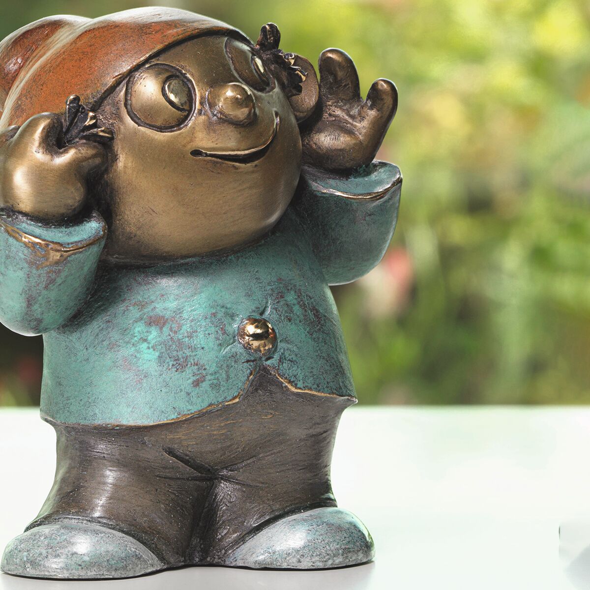 Detailreiche, handpatinierte Bronze-Skulptur eines Mainzelmännchens auf hellem Untergrund mit Garten im Hintergrund. Die Figur trägt eine türkisfarbene Jacke, schwarze Hose und Mütze. Runde Brille, freundlicher Gesichtsausdruck, erhobene Hände mit ausgestreckten Fingern.