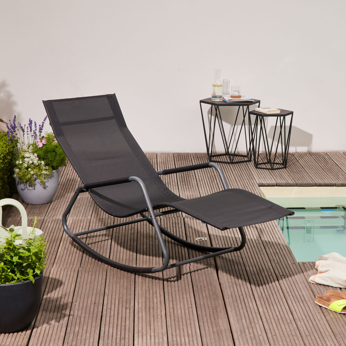 Eine moderne Schaukel-Liege in schwarzer Farbe steht auf einer Holzterrasse neben einem Pool. Umgeben von Pflanzen, einem kleinen Beistelltisch und einer Gießkanne, vermittelt die Szene einen entspannten Outdoor-Lifestyle.