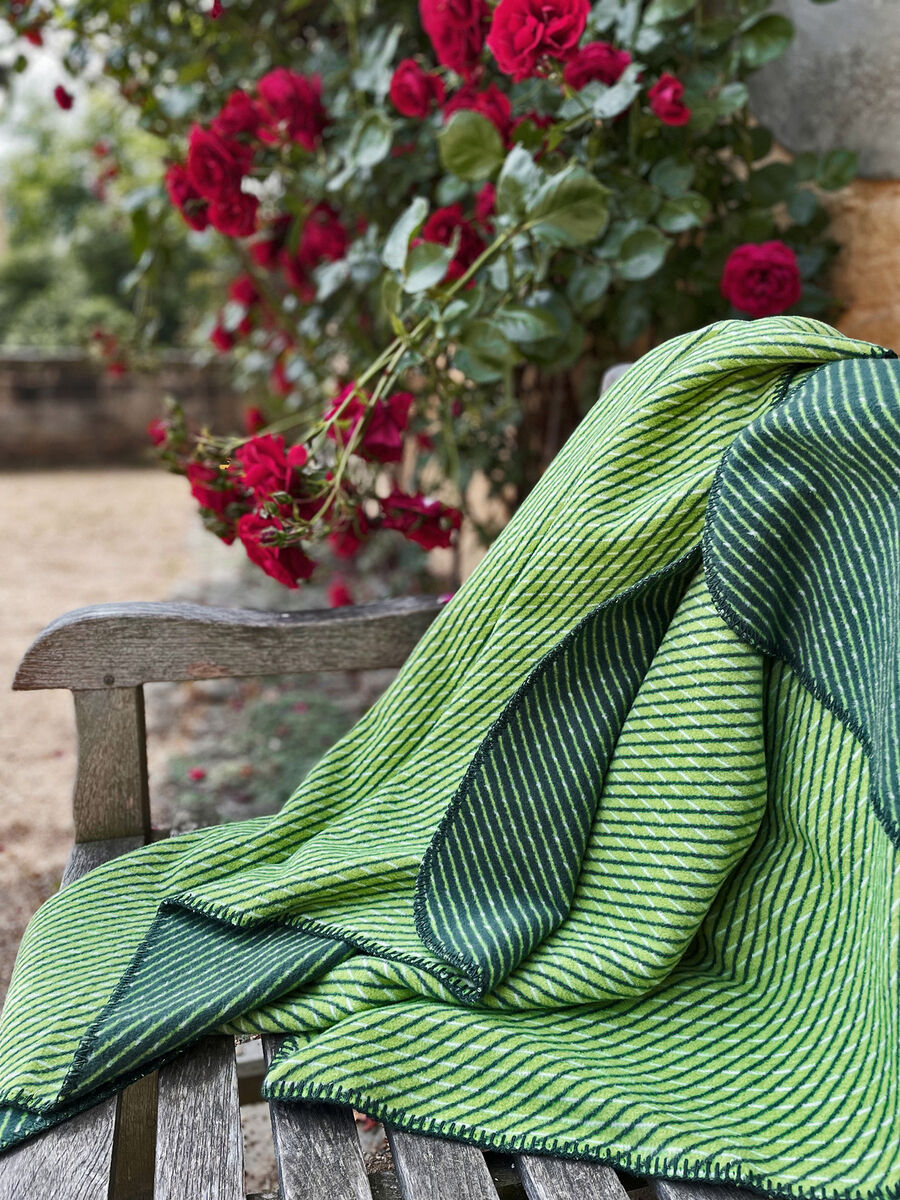 Grüne und dunkelgrüne gestreifte Baumwolldecke »Diagonale« von CORBEZZOLI!® auf einer Gartenbank, mit roten Rosen im Hintergrund.