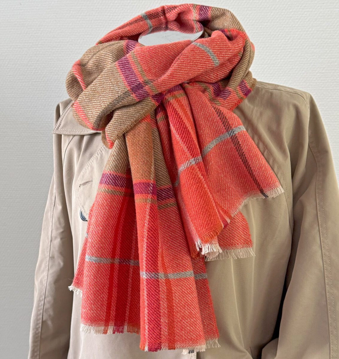 Merinoschal 'Gregor' aus kariertem Wollstoff in Rot-, Orange- und Beigetönen, dekorativ um einen unifarbenen beigefarbenen Mantel gelegt, vor neutralem Hintergrund fotografiert. Ideal als modisches Accessoire für kalte Tage.