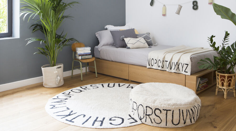 Kinderzimmer mit grauer Wand, Holzbett mit grauer Bettwäsche und Kissen, runder Teppich und Sitzpouf mit Buchstabenmuster