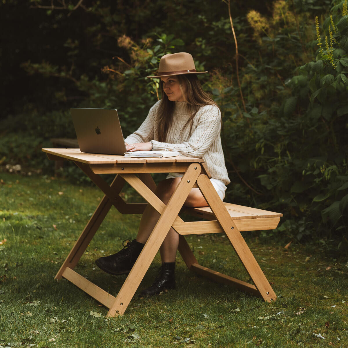 Eine Frau sitzt im Garten an einer klappbaren Tischbank „Folding Picnic Table“ von Weltevree aus braunem Bambus. Sie arbeitet mit einem Laptop, trägt Hut, Strickpullover und Boots. Die Szene ist von grüner Natur umgeben und vermittelt eine entspannte Outdoor-Atmosphäre.