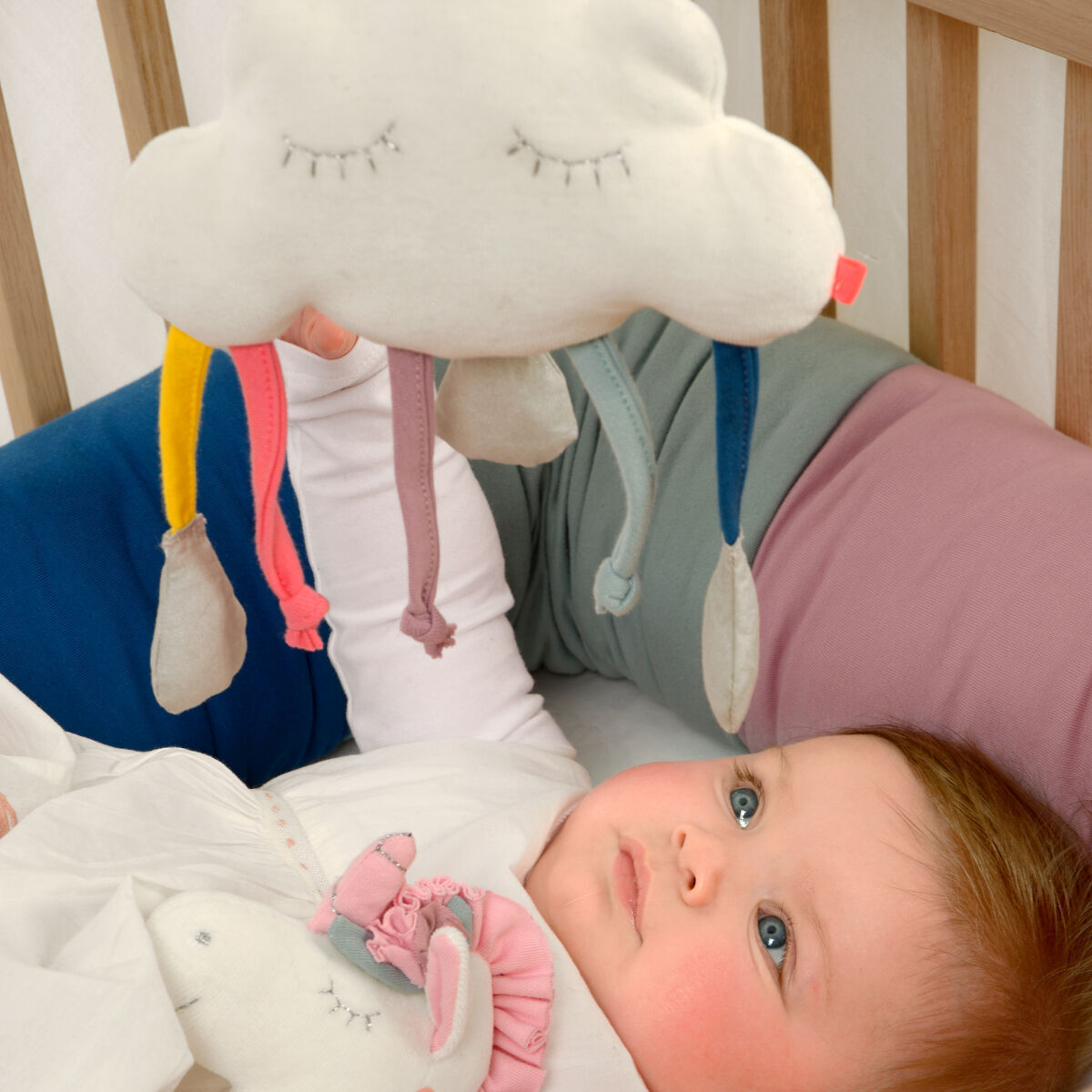 Ein Baby liegt in einem Kinderbett und schaut auf eine wolkenförmige Spieluhr mit bunten Bändern, die darüber hängt.