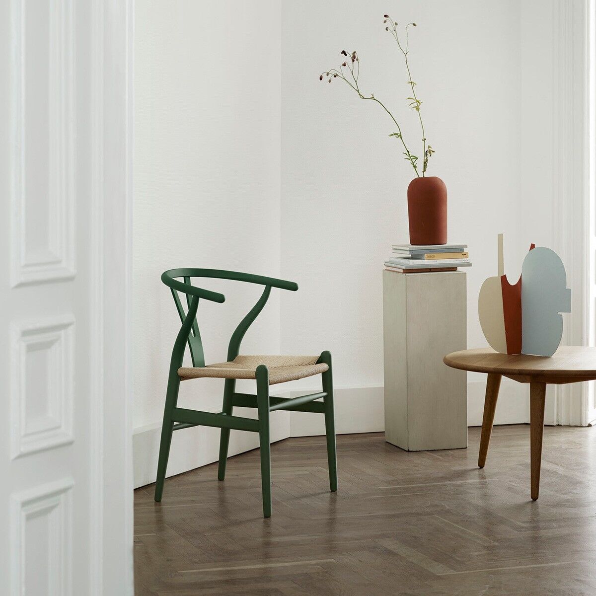 Ein Designstuhl CH24 Soft von Carl Hansen steht in einem stilvollen Wohnzimmer auf einem Holzboden neben einem minimalistischen Sockel mit Dekoration und einer Vase. Der Stuhl hat eine grüne Lackierung und eine beige Sitzfläche.