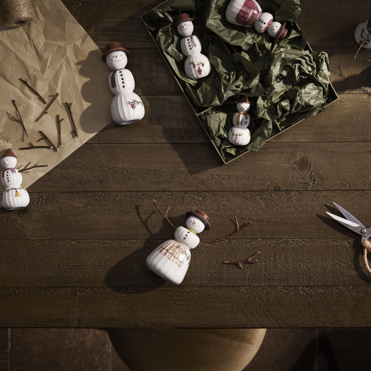 Verschiedene Schneefrau-Keramikfiguren der Serie »Hammershøi« liegen und stehen auf einem hölzernen Tisch, umgeben von Bastelmaterialien wie Zweigen, Schere, Draht und Papier in Naturtönen sowie einer Tasse mit orangefarbenem Getränk. Die Schneefrauen tragen braune Hüte und dekorative rote Details.