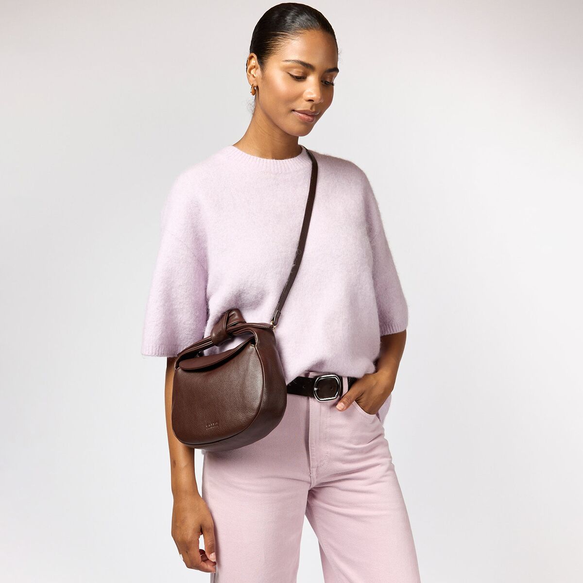 Eine Frau trägt die Tasche »Coco Crossiant« von O My Bag aus luxuriösem Soft-Narbenleder in Schokoladenbraun diagonal über ihre Schulter. Sie trägt einen weichen hellrosa Pullover, eine hellrosa Hose mit schwarzem Gürtel. Die Tasche hat einen abgerundeten, kompakten Look mit glatter Oberfläche und dezenter Prägung.