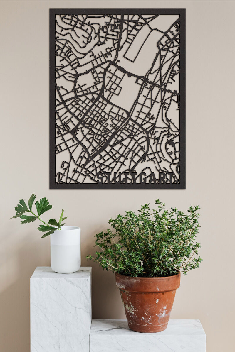 Eine schwarze Holzstadtkarte von Stuttgart hängt an einer beigen Wand. Darunter stehen auf einem weißen, marmorierten Sockel eine grüne Topfpflanze und eine weiße Kerze.