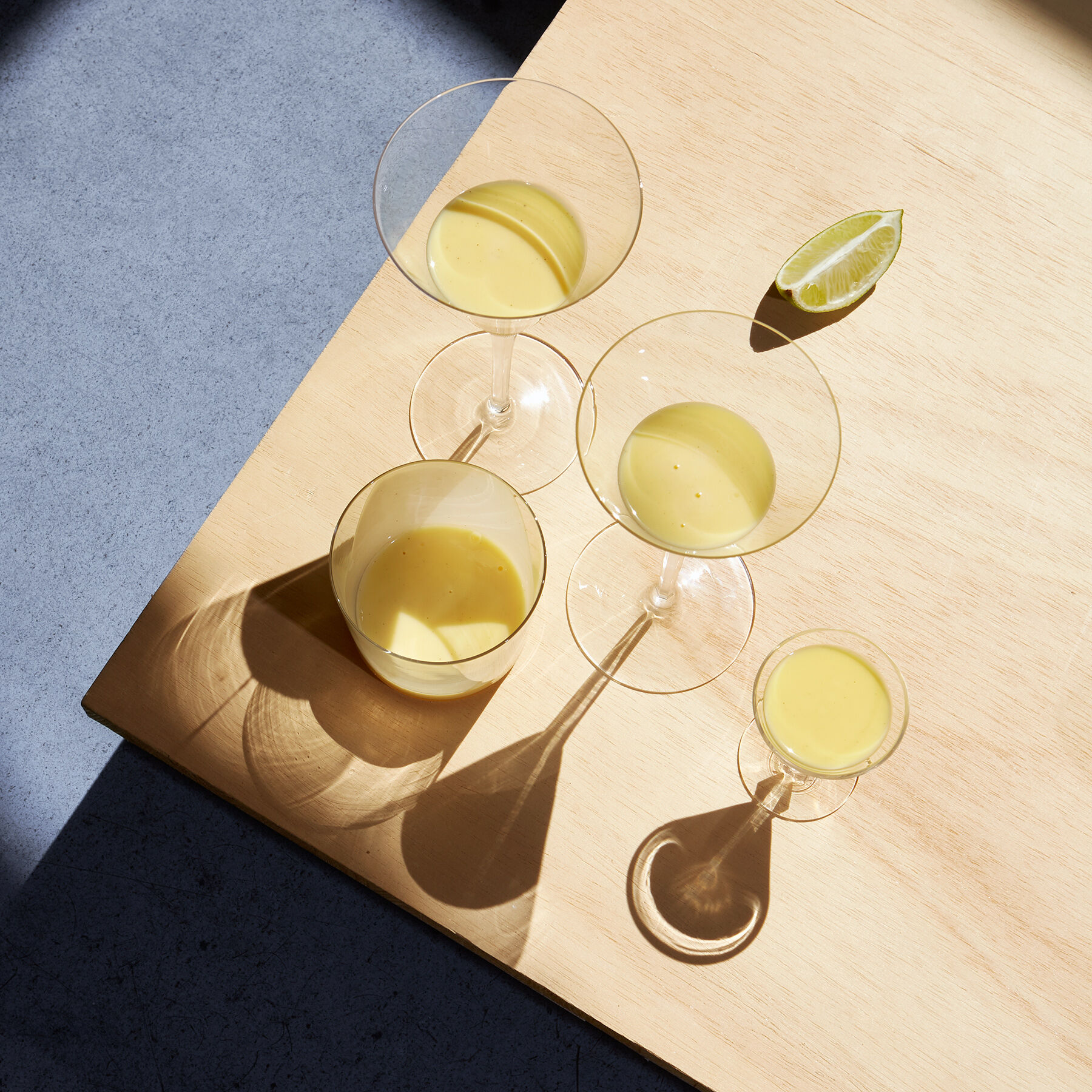Ein Arrangement von mehreren Gläsern, die mit einem cremigen, gelben Eierlikör gefüllt sind, steht auf einem Tisch aus hellem Holz. Ein Limettenschnitz liegt ebenfalls auf dem Tisch, und Schatten von Gläsern fallen auf die Oberfläche.