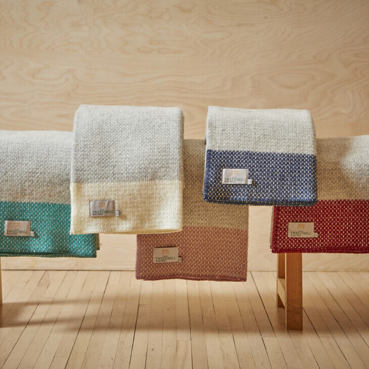 Eine Holzbänk mit vier gefalteten Decken der Marke 'Crossweave'. Jede Decke hat eine zweifarbige Strickmusterung in den Farben Beige-Grau, Türkis-Weiß, Rosa-Grau und Rot-Weiß. Die Decken hängen nebeneinander auf der Bank.