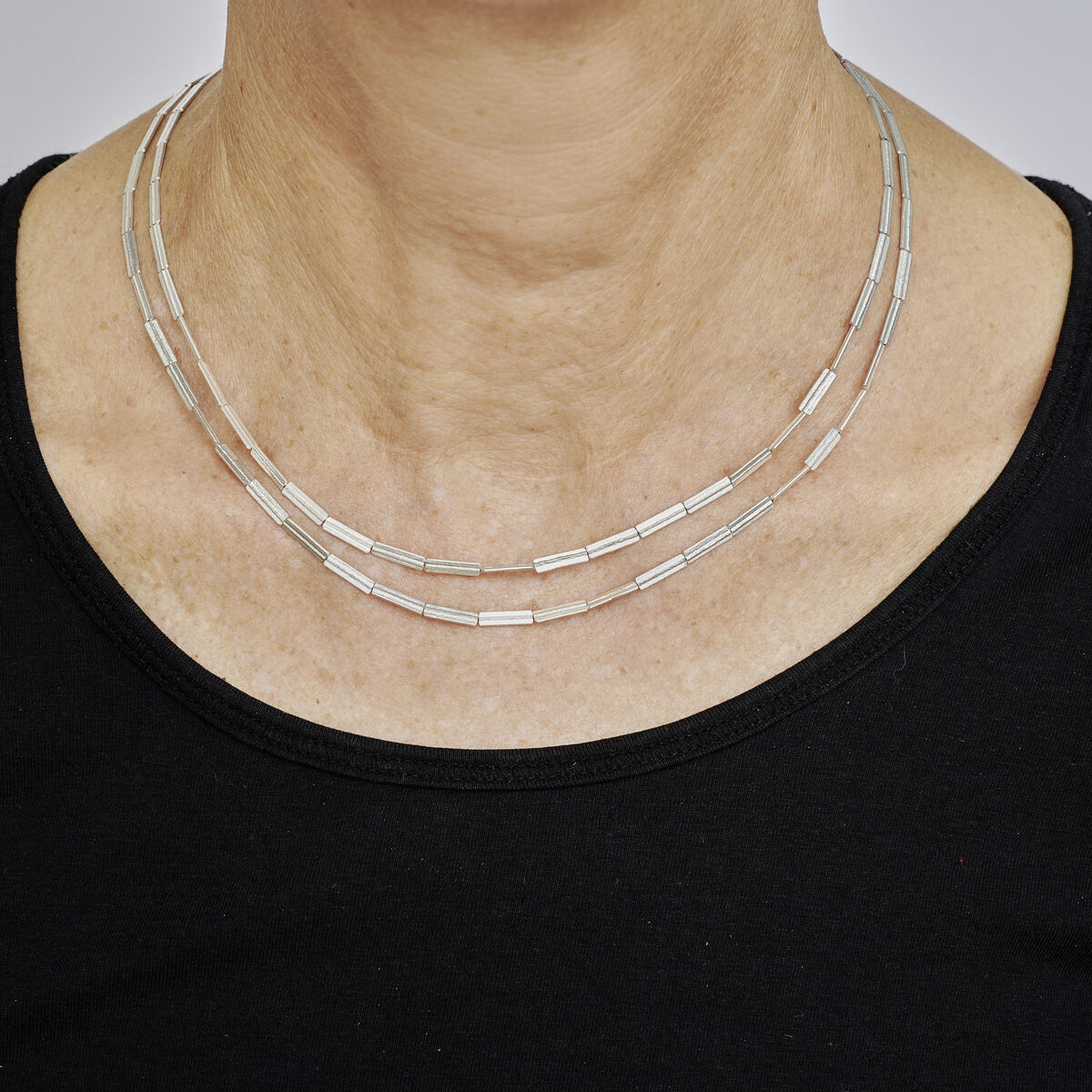 Nahaufnahme eines zweireihigen Colliers aus geprägtem Silber, das um den Hals einer Person über einem schwarzen Oberteil getragen wird. Das Schmuckstück wirkt modern und elegant, mit rechteckigen Gliedern und glänzender Oberfläche, fotografiert vor einem neutralen Hintergrund.