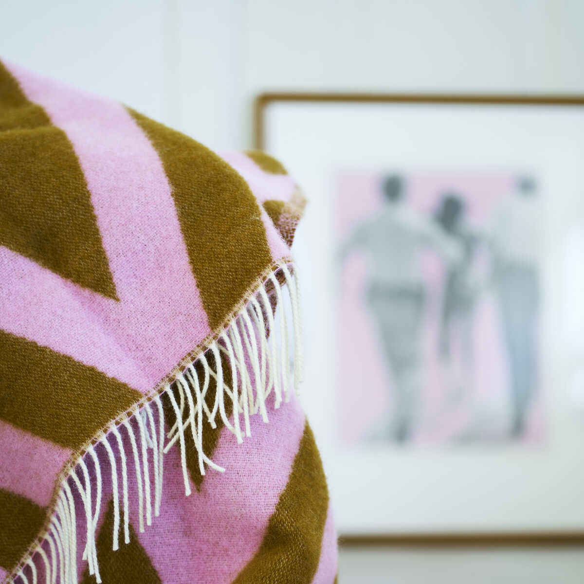 Weiche Decke mit Zickzack-Muster in Rosa und Braun, dekorativ auf einer hellen Oberfläche drapiert, im Hintergrund ein gerahmtes Bild mit verschwommenem Motiv.