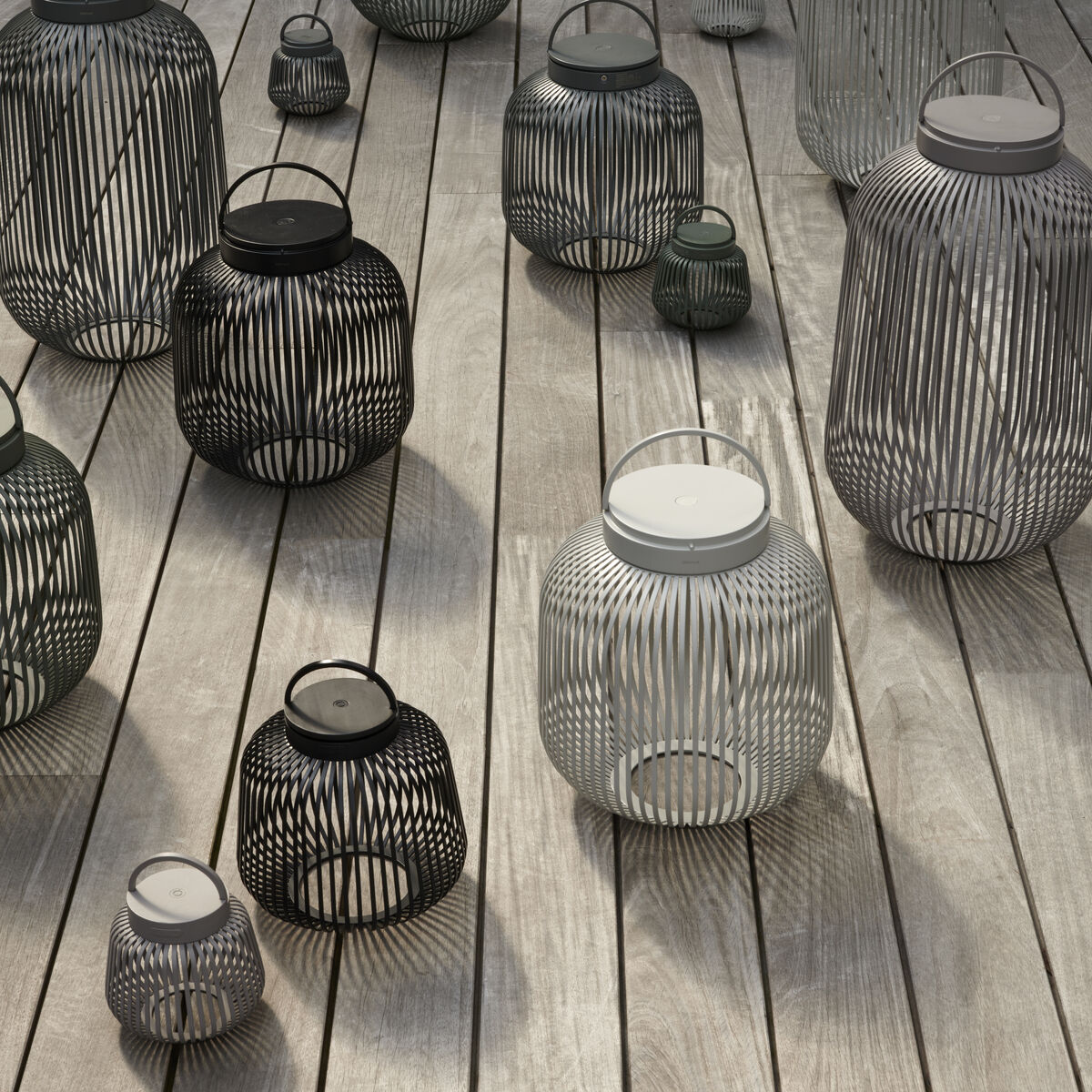 Verschiedene LED-Leuchten 'LITO' von blomus in schwarzem, grauem und weißem Metall stehen stilvoll auf hellen Holzdielen. Die rundlichen, offenen Metallkörper erzeugen ein interessantes Schattenspiel, ideal für moderne Terrasse oder Garten.