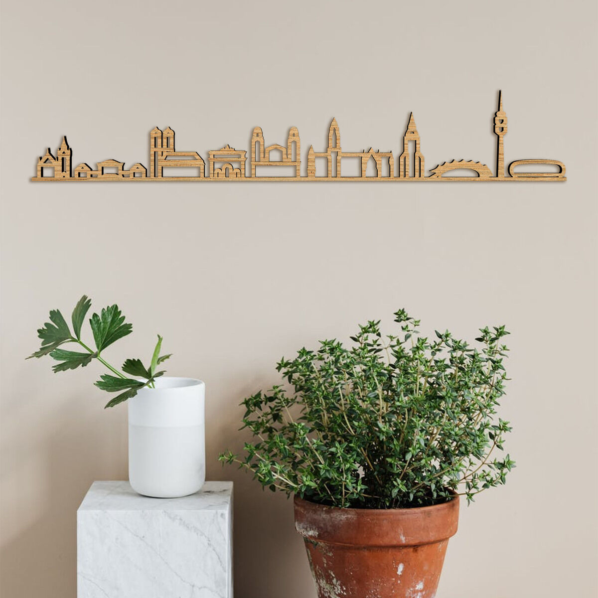 Eine Holzkunst-Silhouette der Skyline von München, die dekorativ an einer beige-farbenen Wand hängt. Daneben stehen ein weißer Blumentopf und ein Terrakottatopf mit grünen Pflanzen auf einem Marmorpodest.
