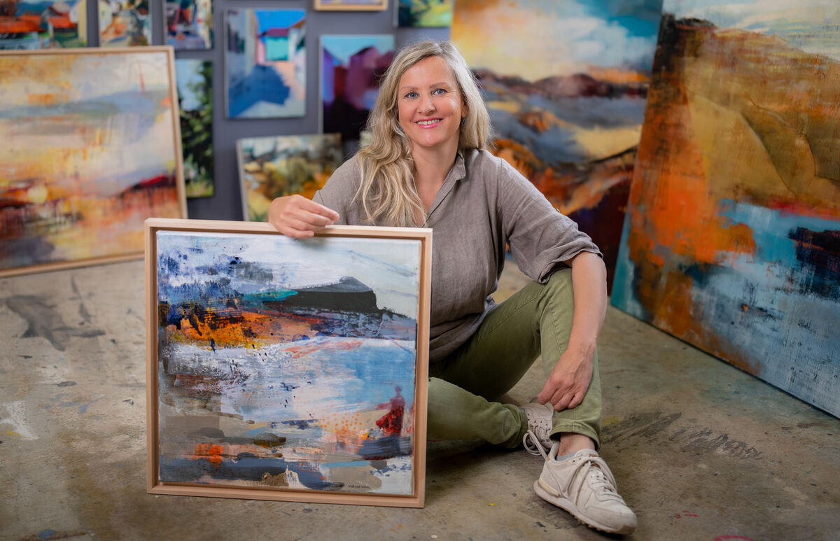 Eine Frau, die auf dem Boden eines Ateliers sitzt und ein abstraktes Gemälde hält. An den Wänden sind weitere Kunstwerke zu sehen. Die Künstlerin lächelt und trägt eine legere Kleidung. Das Atelier ist mit bunten, abstrakten Gemälden dekoriert.