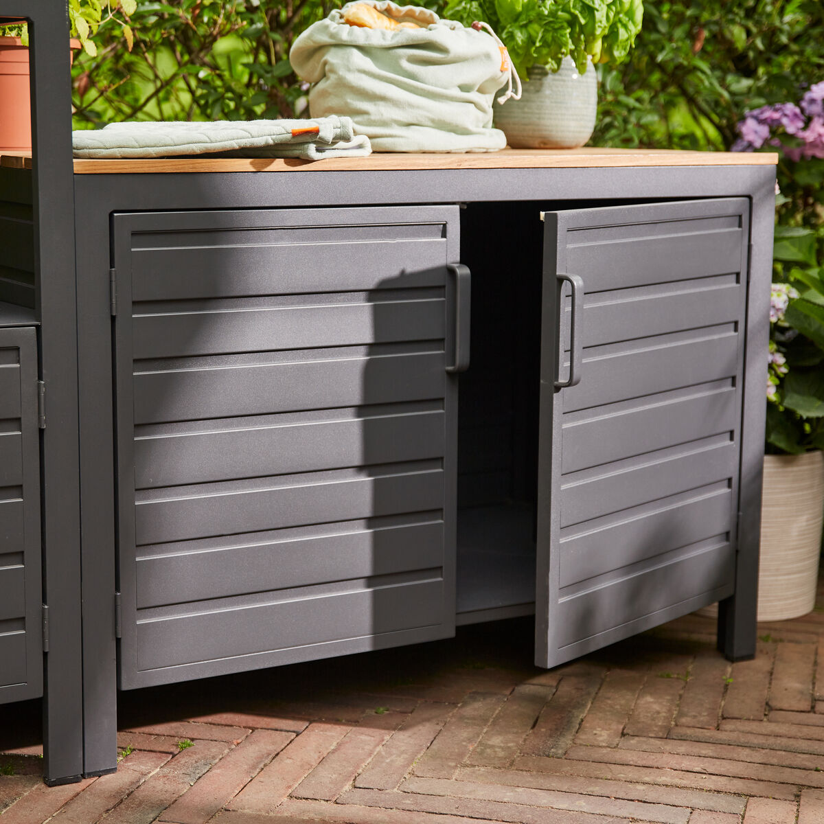 Outdoor-Küchenmodul mit einer grauen, wetterbeständigen Struktur und einer natürlichen Holzplatte, flankiert von Pflanzen in einem Gartenbereich.