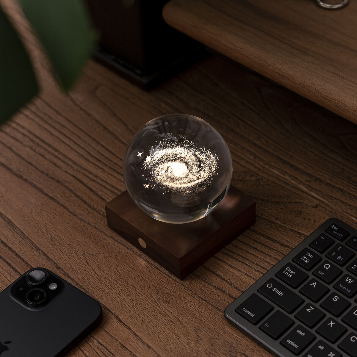 Kleine Tischlampe namens 'Amber Light' mit runder Glaskugel und leuchtendem Zentrum auf einer eckigen Holzbasis, auf einem Holzschreibtisch neben Tastatur, Smartphone und Schlüsselanhänger platziert.