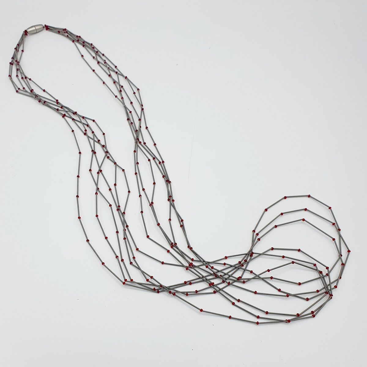 Eine lange, mehrsträngige Kette aus dünnen Metallstäben, verbunden durch rote Perlen, auf weißem Hintergrund.