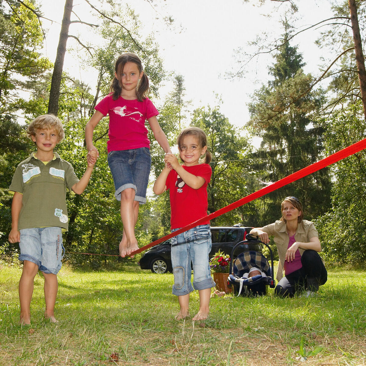 Kinder balancieren auf einer roten Slackline im Freien, während ein Elternteil im Hintergrund sitzt. Die Slackline ist zwischen Bäumen gespannt. Die Szene spielt sich in einer grünen, bewaldeten Umgebung bei sonnigem Wetter ab.