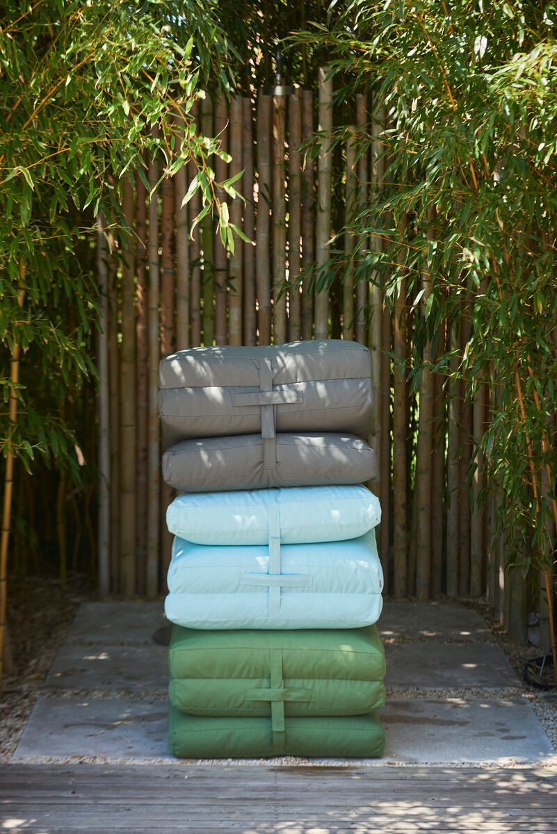 Eine gestapelte Gruppe von drei Gartenmatratzen in den Farben Grau, Blau und Grün steht in einem Außenbereich vor einem Hintergrund aus Bambus.