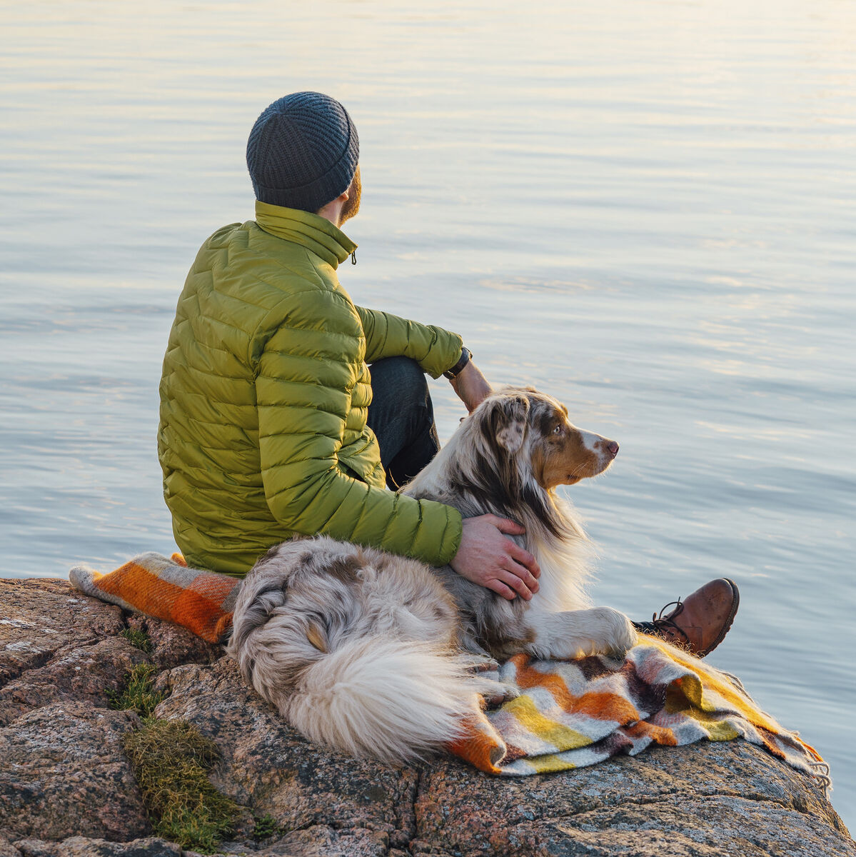 Eine Person in einer grünen Jacke sitzt neben einem Hund auf einem Felsen am Wasser. Beide blicken auf den See, während sie auf einer bunten Decke sitzen. Die Szene strahlt Ruhe und Gelassenheit aus.