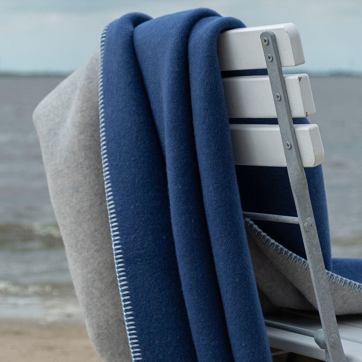 Eine weiche Merino-Decke in dunklem Blau und dezentem Hellgrau liegt über einer weißen Holzlehne eines Stuhls am Strand mit Blick auf das Meer. Die Szene wirkt ruhig und einladend, ideal für entspannte Momente im Freien oder zum Kuscheln im Haus.