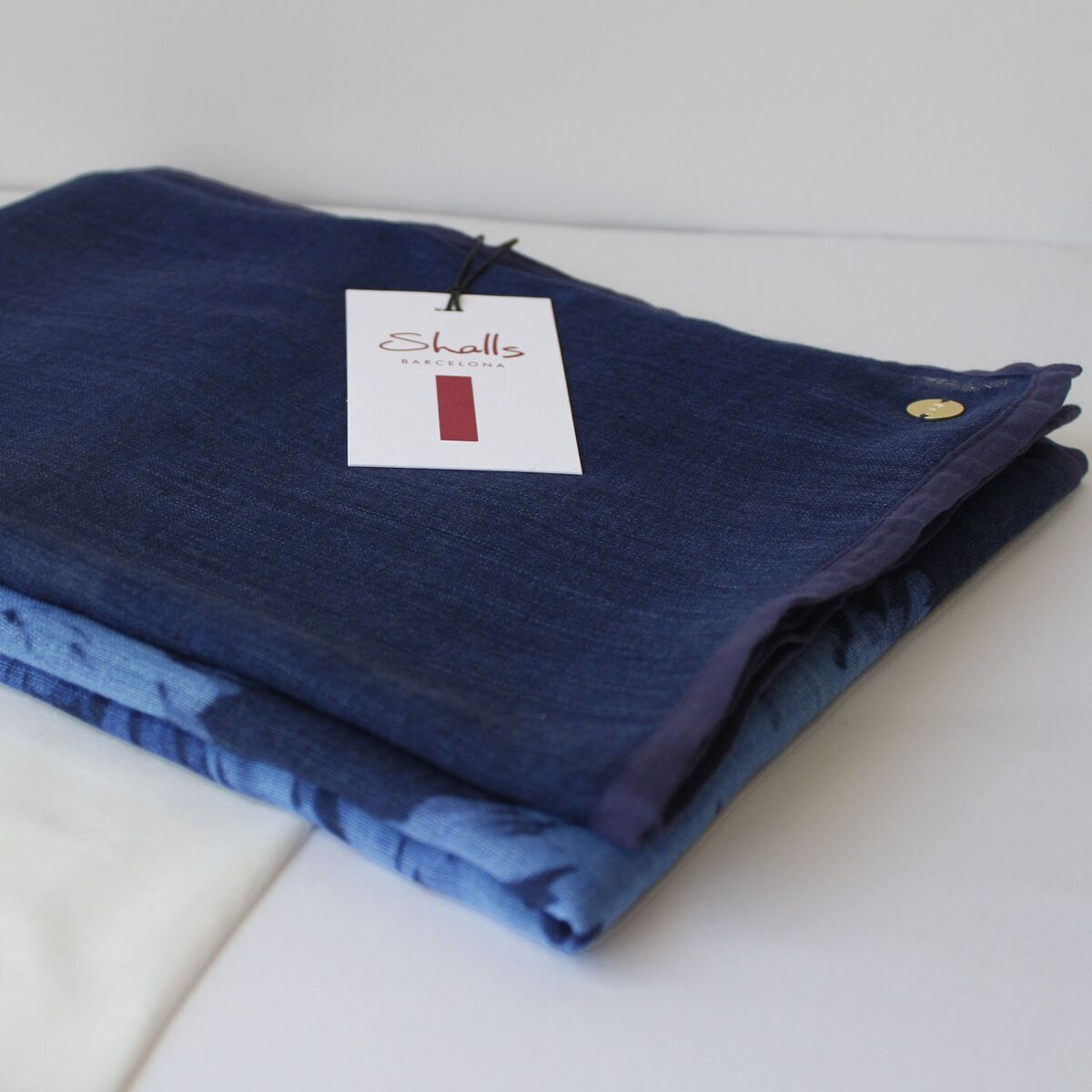 Ein zusammengefalteter, dunkelblauer Wollschal liegt auf einem hellblauen Stoff auf weißem Untergrund. Ein Produktanhänger mit Schriftzug ist am Schal befestigt. Daneben befindet sich ein weißer Stoffbeutel.