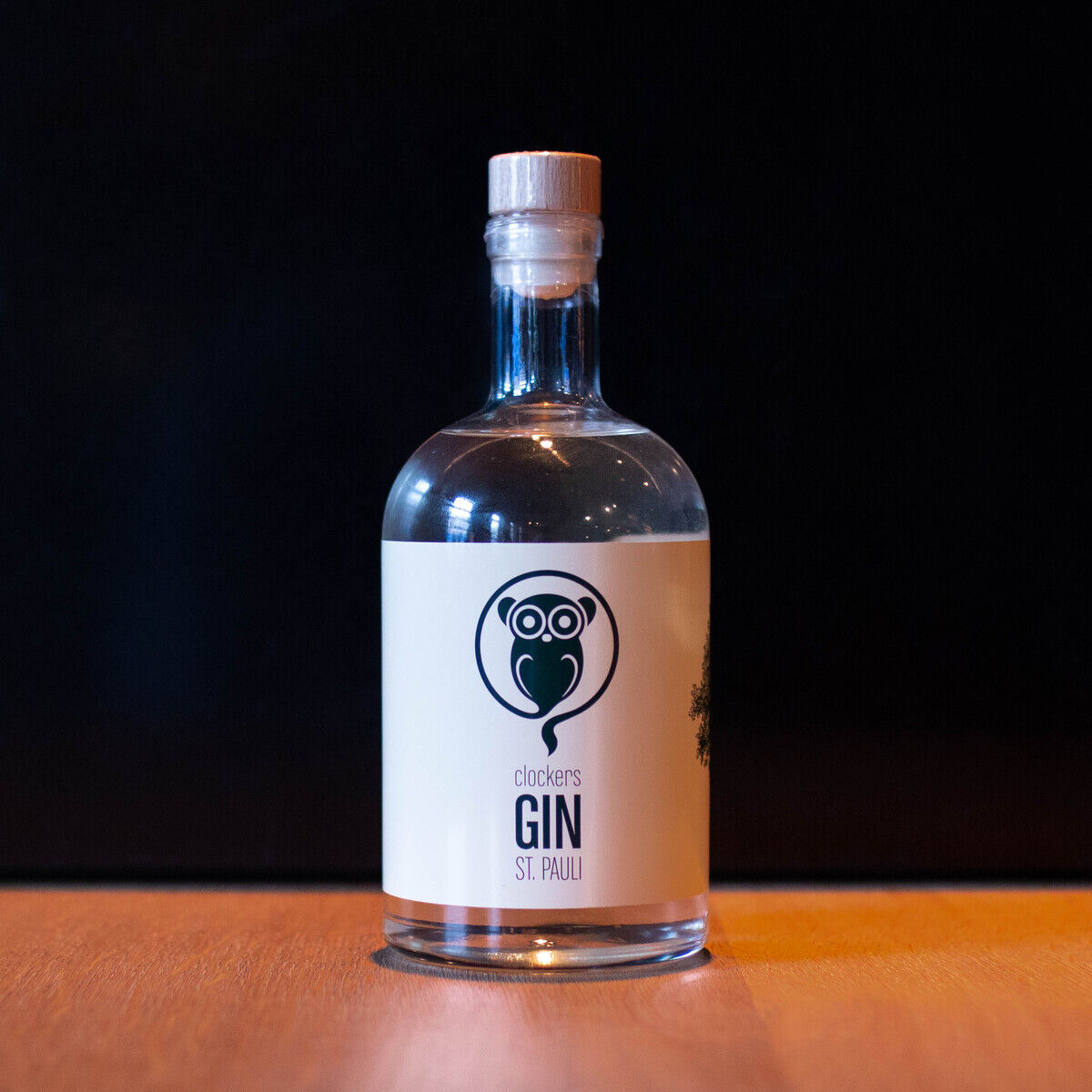 Eine Flasche clockers Gin St. Pauli steht auf einem Holzuntergrund vor einem dunklen Hintergrund. Das Etikett der Flasche hat ein Eulendesign und die Farben sind klar, weiß, schwarz und braun.
