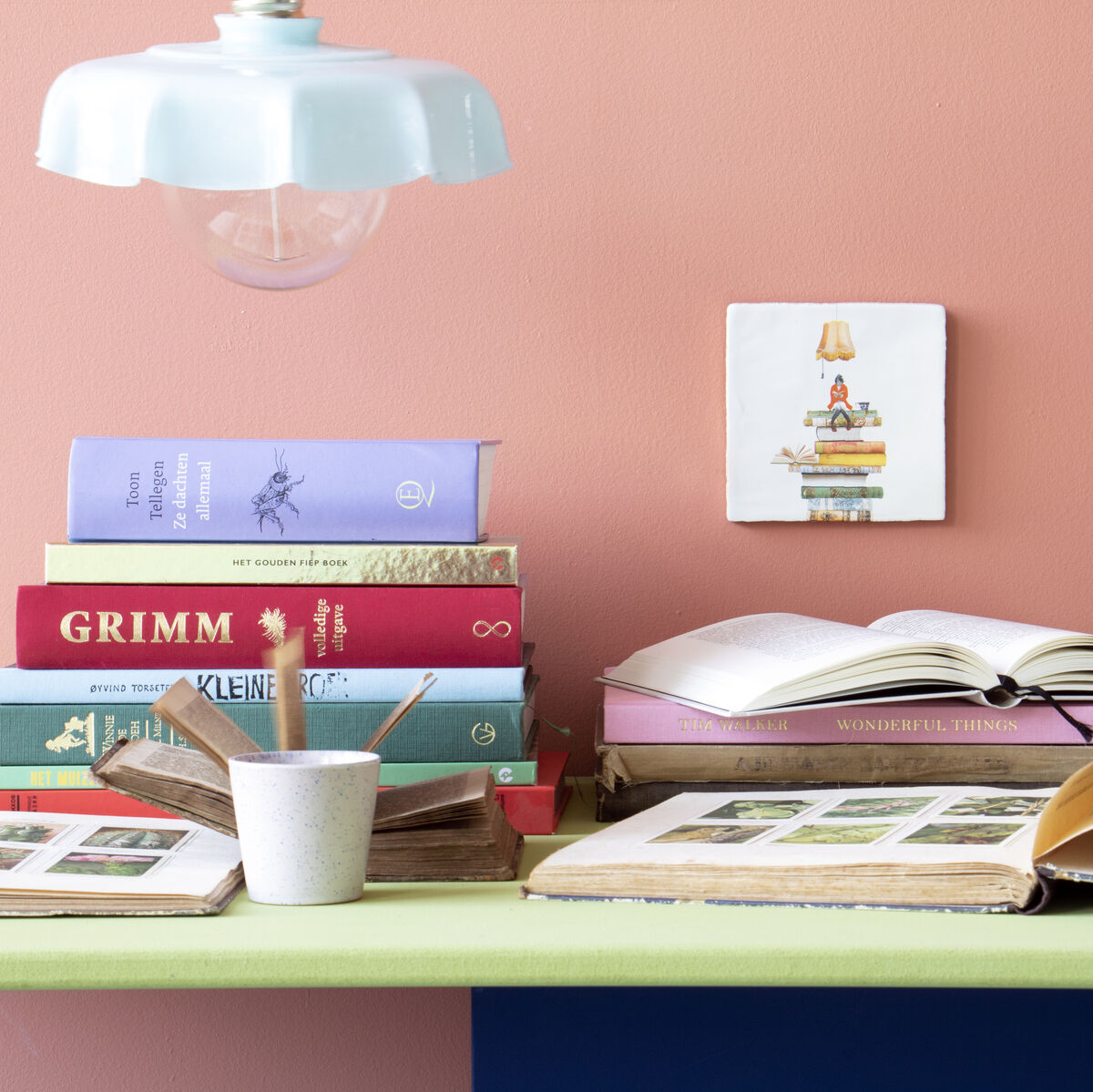 Eine Szene auf einem Schreibtisch vor einer rosa Wand. Mehrere Bücher sind gestapelt, dekoriert mit einer Keramikfliese, die ein »Bücherwurm«-Motiv zeigt. Eine Lampe hängt über dem Schreibtisch, und ein Becher mit Stiften steht daneben.