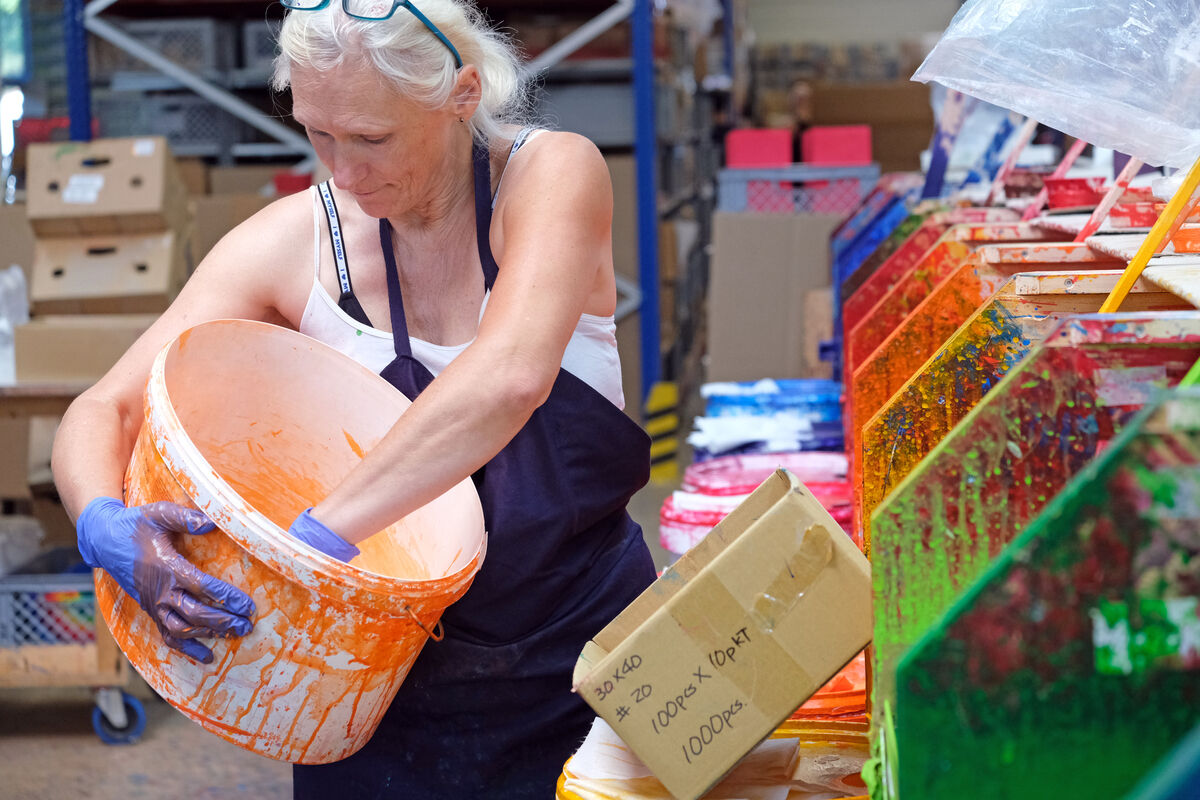 Eine ältere Frau in einer Werkstatt trägt blaue Handschuhe und ein dunkles Schürzenkleid mit weißen Trägern, während sie in einen großen orangefarbenen Eimer greift, der vor einer Reihe bunter Plexiglastrichter steht. Kartons und verschiedene Farben füllen den Hintergrund.