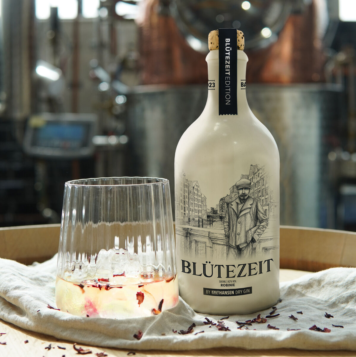 Eine Flasche ZEIT-Jubiläumsedition Gin »BLÜTEZEIT« steht neben einem Glas mit Cocktail, dekoriert mit Blütenblättern, auf einem Holztisch in einer Destillerie.