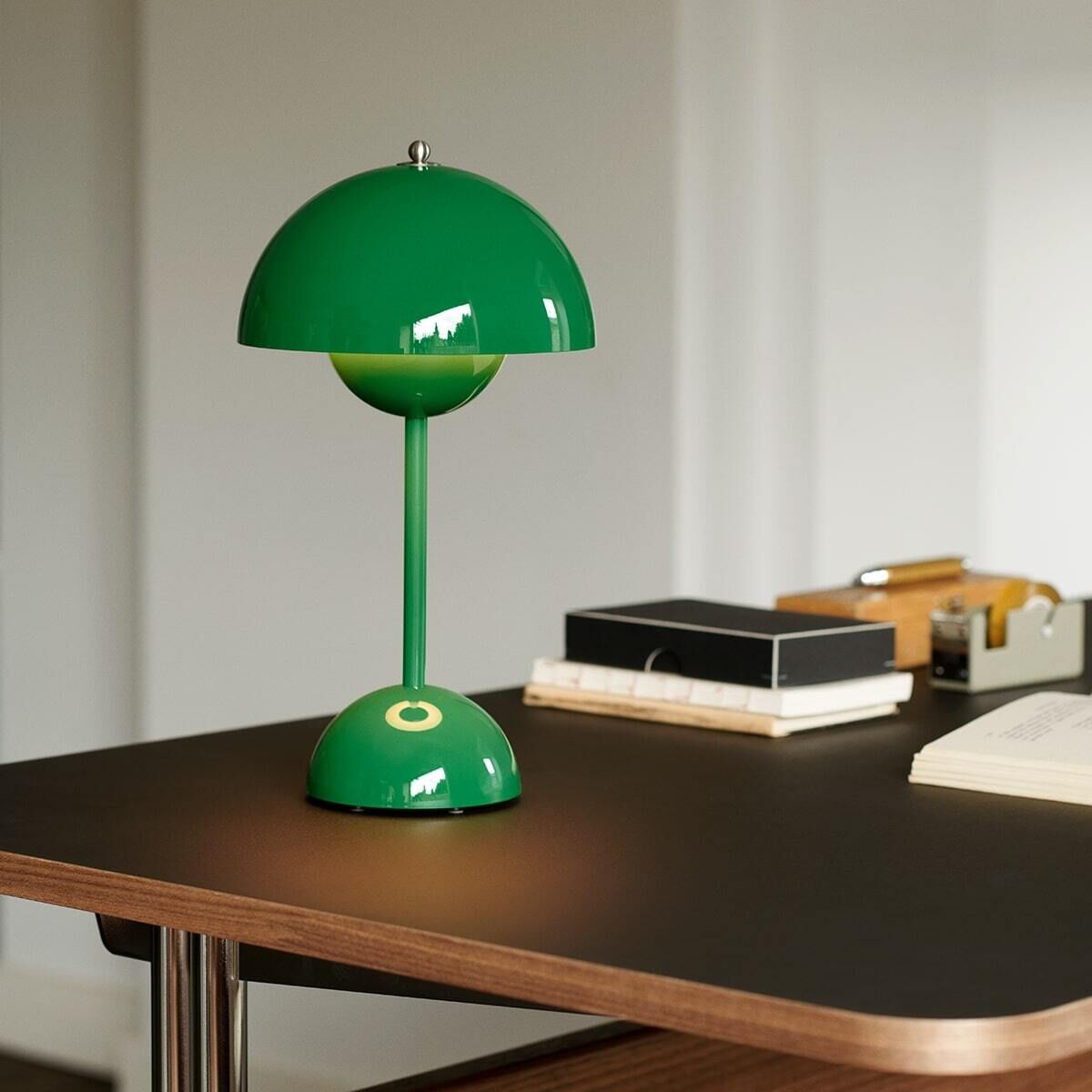Die Flowerpot VP9 Tischleuchte in Akkubetrieb steht auf einem dunklen Holztisch in einem modern eingerichteten Innenraum. Die Lampe hat ein auffallend grünes, halbrundes Design mit einem schmalen Ständer und einem dekorativen Schirm. Im Hintergrund sind ein paar Bücher und einige Büroartikel auf dem Tisch zu sehen.