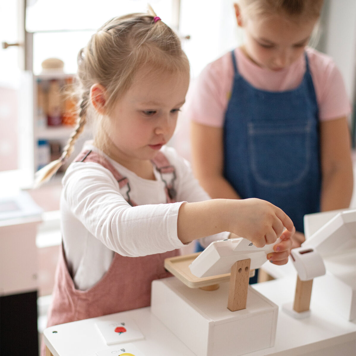 Zwei Kinder spielen mit einer Holz-Waage in einem Indoor-Spielbereich. Die Waage befindet sich auf einem kleinen weißen Tisch und ist Teil eines Marktstand- oder Küchenrollenspiels.