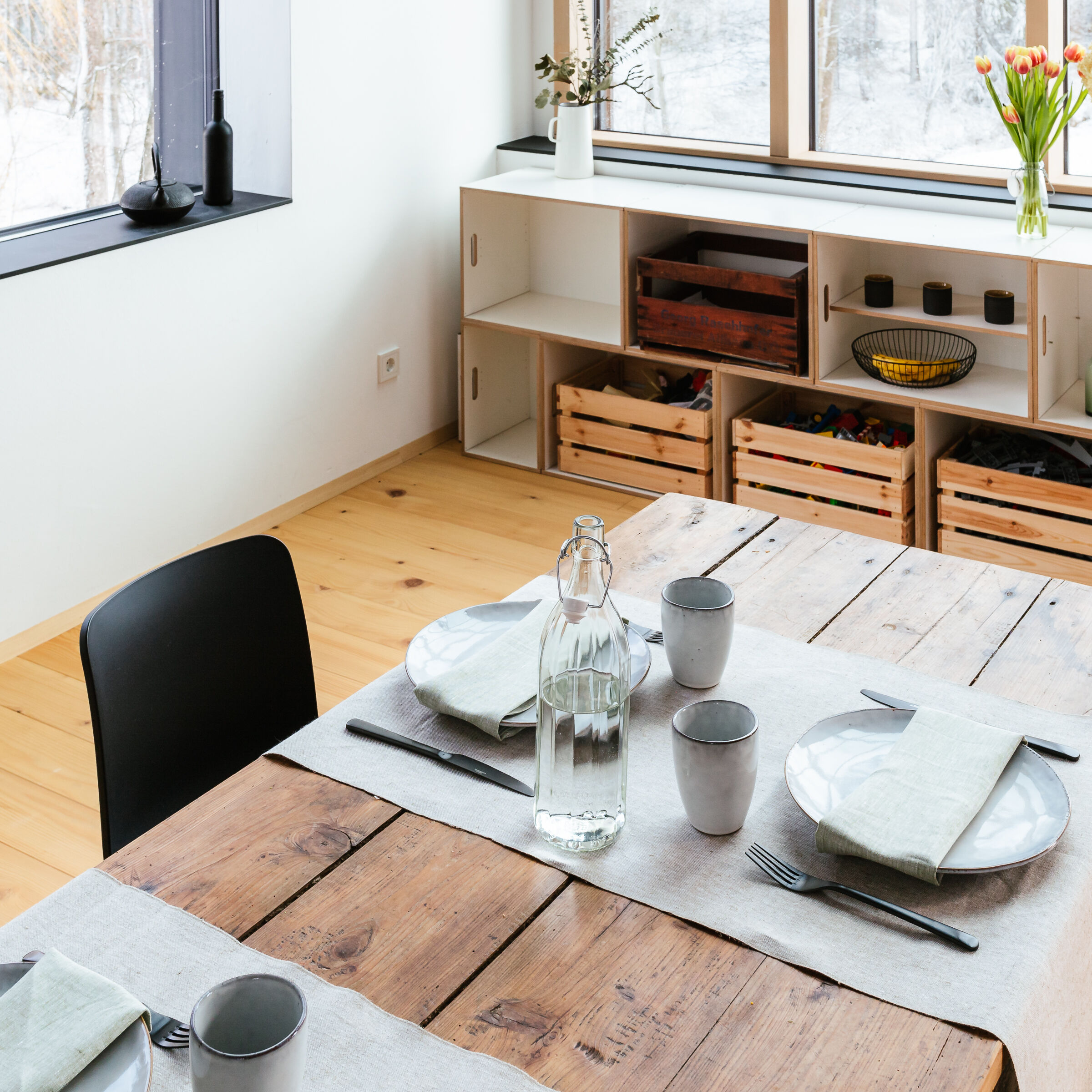 Ein modernes Esszimmer mit skandinavischem Stil. Auf einem Holztisch liegt ein Leinen-Tischläufer. Der Tisch ist mit Geschirr und einer Wasserkaraffe gedeckt. Im Hintergrund sind Holzmöbel mit dekorativen Elementen und eine Vase mit Blumen.