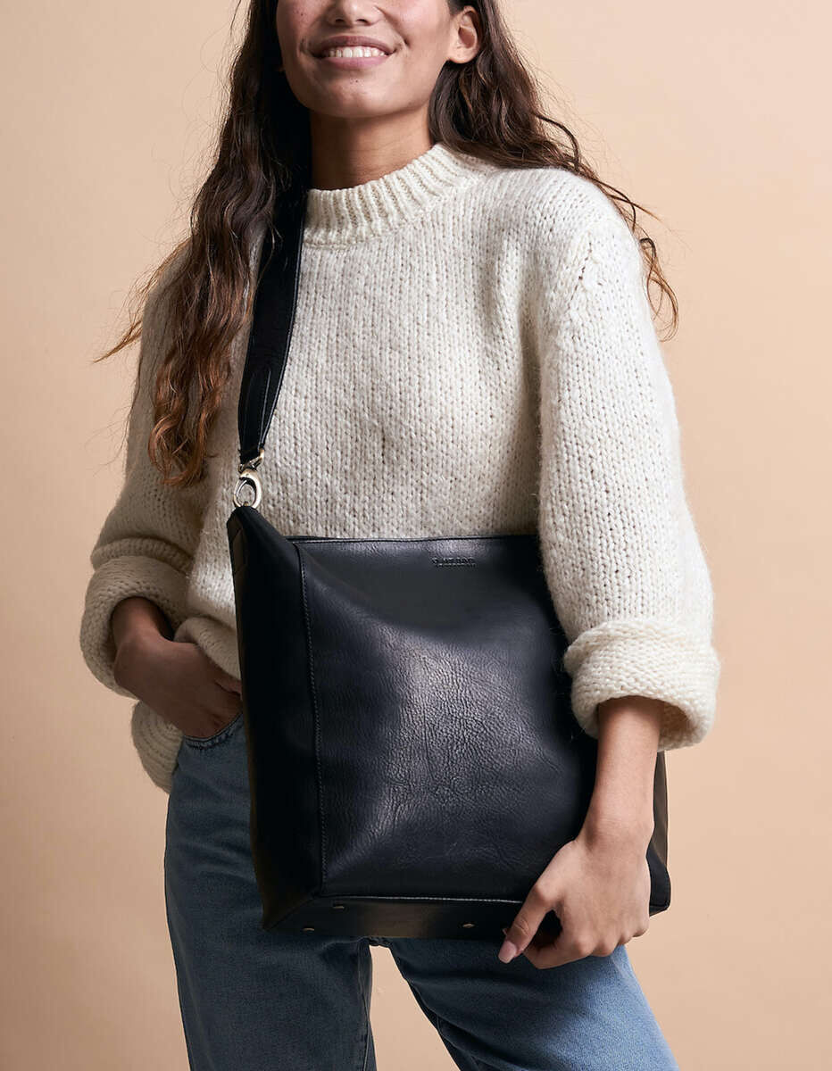 Eine Frau trägt eine schwarze Ledertasche »Sofia« über ihrer Schulter. Sie hat einen hellen Pullover und eine hellblaue Jeans an und steht vor einem beigen Hintergrund.