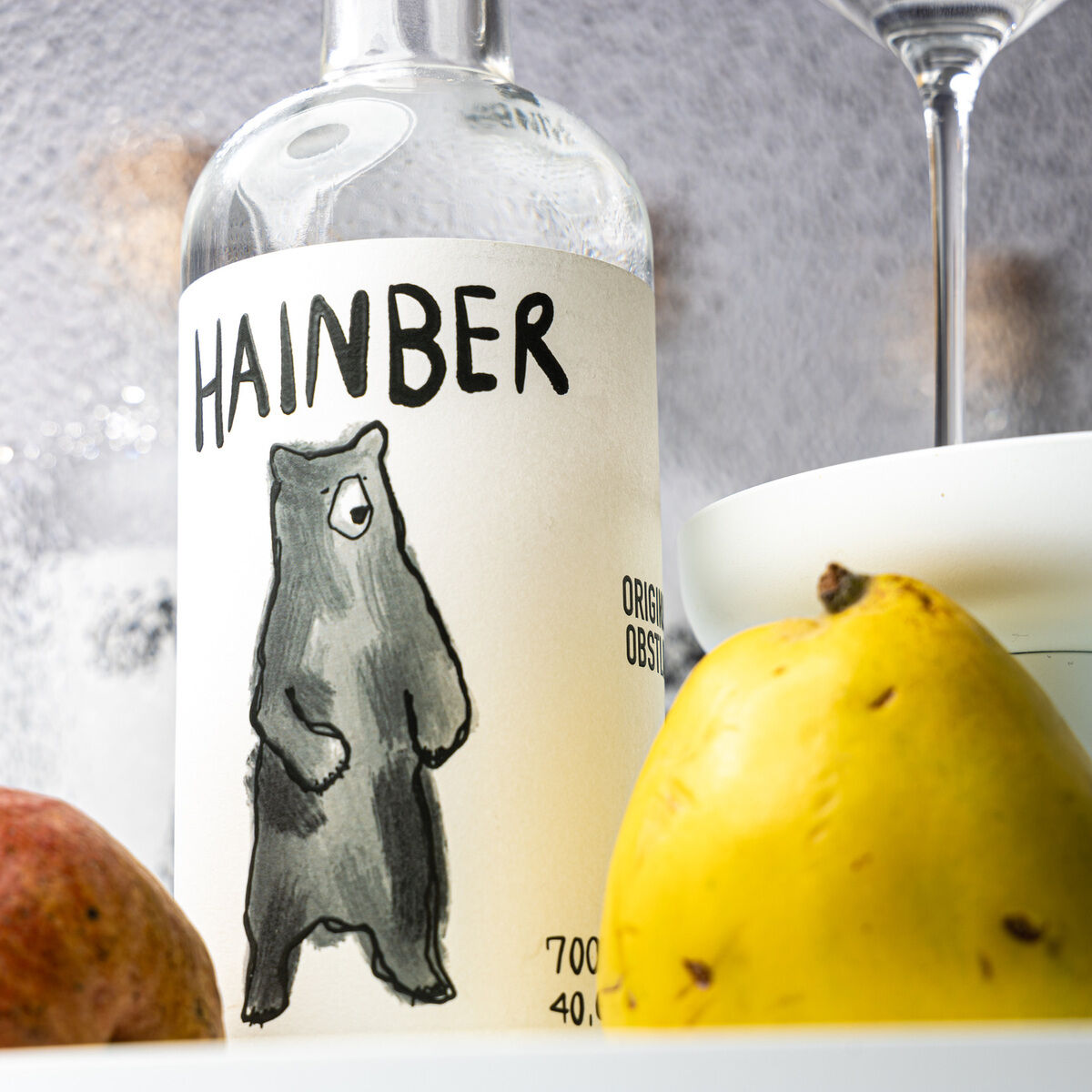 Im Vordergrund stehen eine Flasche Marillen-Obstler "HAINBER" von AASPIRITS mit gezeichnetem Bären auf dem Etikett, eine gelbe Birne und eine weitere Frucht. Im Hintergrund sind ein Weißweinglas und ein Glas mit goldener Flüssigkeit zu sehen. Helle, moderne Atmosphäre.