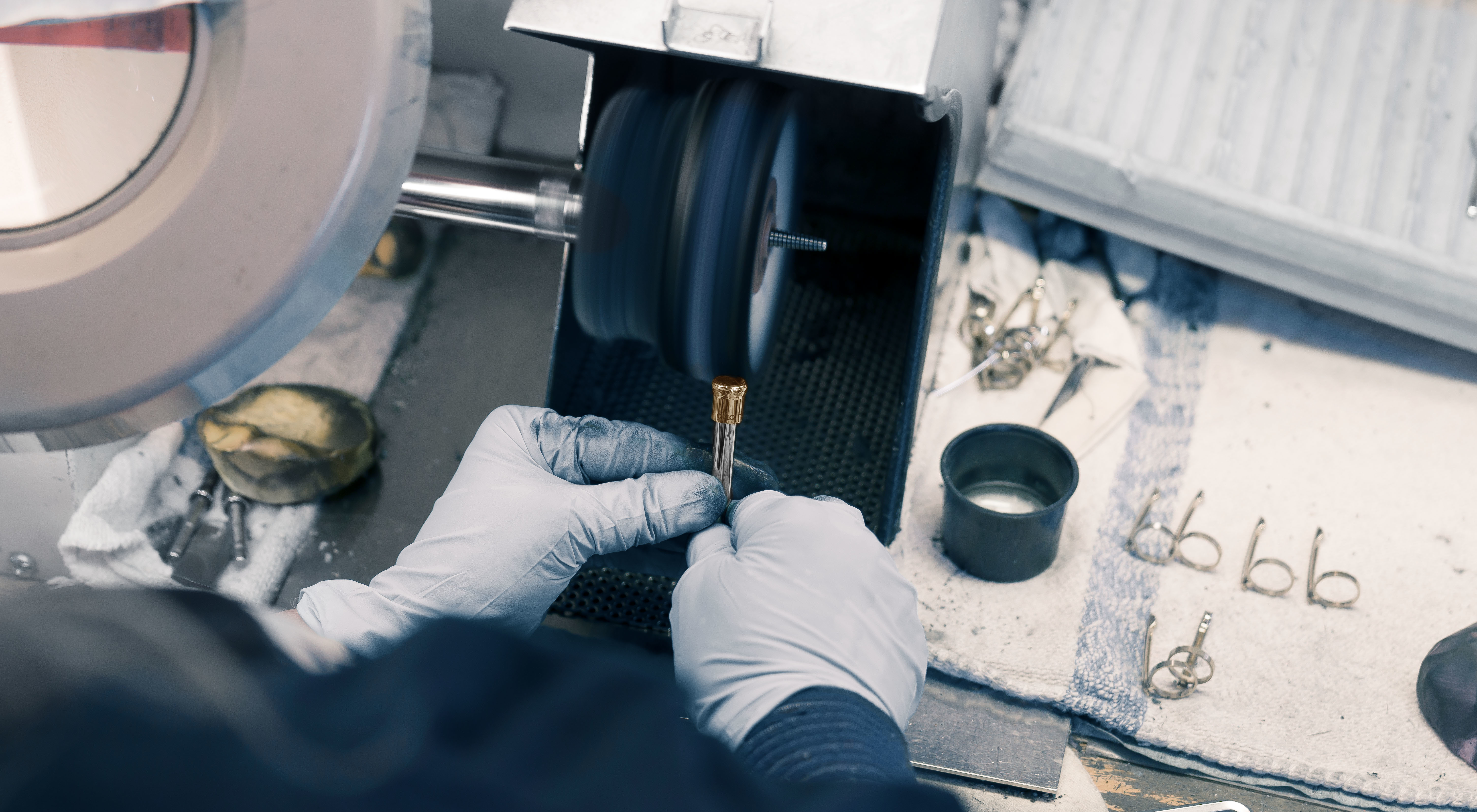 Person mit Handschuhen arbeitet an einem kleinen Metallteil an einer Maschine, umgeben von Werkzeugen und Metallringen