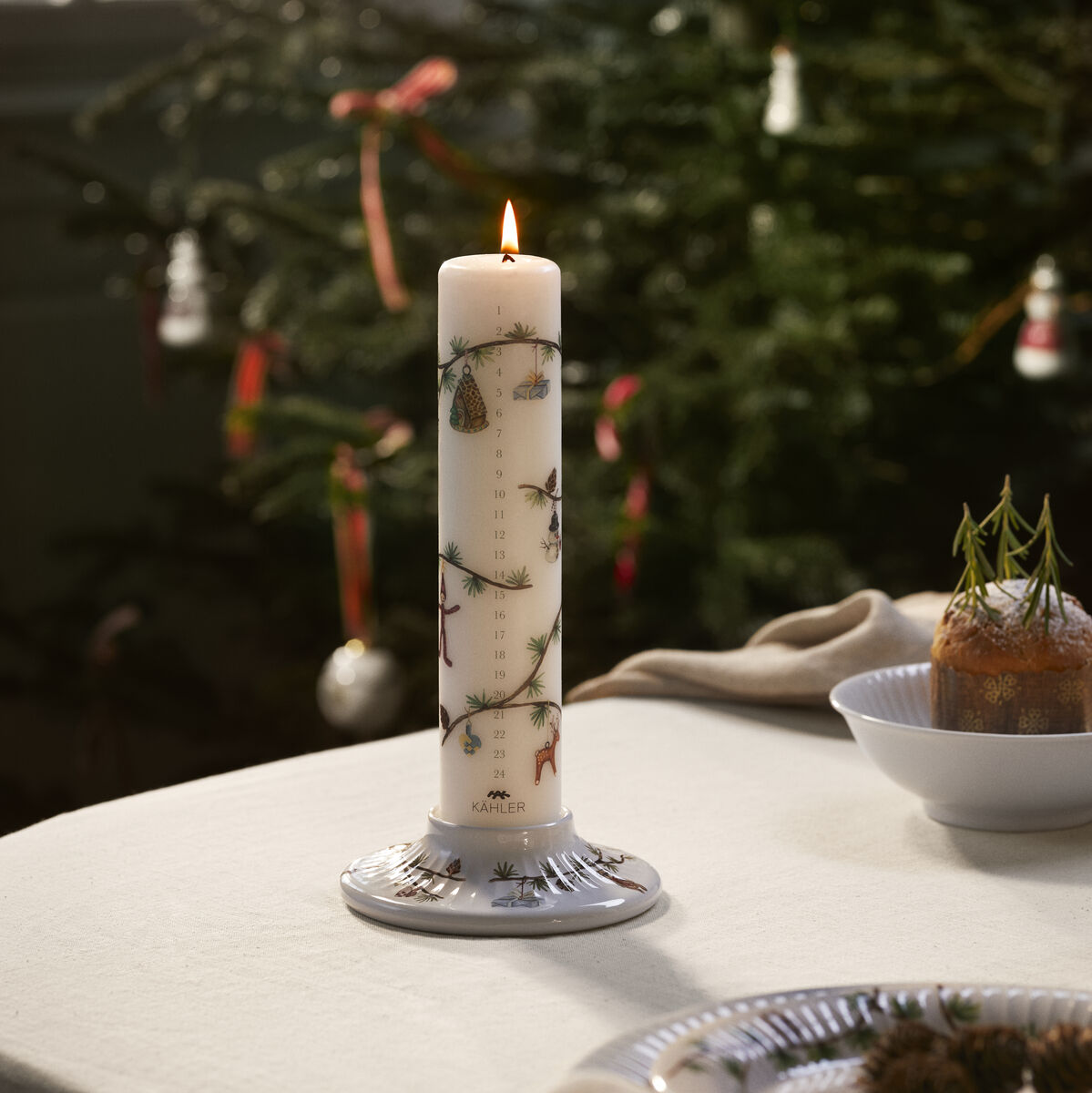 Eine beige Kalenderkerze Hammershøi Ø5 cm steht auf einem festlich gedeckten Tisch vor einem dekorierten Weihnachtsbaum. Die Kerze brennt und verbreitet eine gemütliche Atmosphäre.