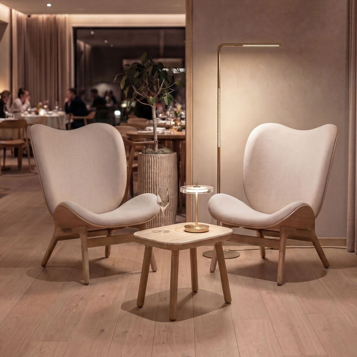 Zwei moderne Lounge-Sessel aus hellem Holz und beige gepolstertem Bezug stehen an einem kleinen, runden Holztisch. Daneben die Stehleuchte »Omni« von Umage in Messing. Der Raum ist warm beleuchtet, die Stimmung ist gemütlich und einladend.