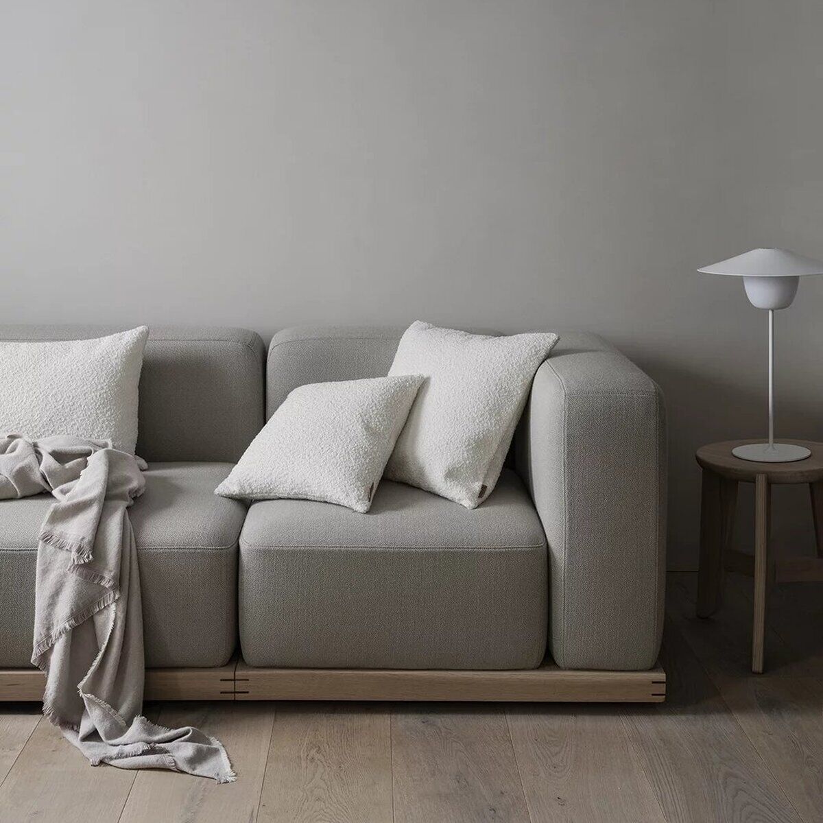 Ein moderner Sitzhocker namens 'ELI' steht in einem skandinavisch eingerichteten Wohnzimmer auf einem hellen Holzboden. Der Sitzhocker ist mit hellgrauem Stoff bezogen und wird von zwei weißen Kissen und einer beigefarbenen Decke ergänzt. Rechts daneben steht ein kleiner Holztisch mit einer schlichten weißen Tischlampe vor einer hellgrauen Wand.