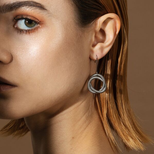 Eine Frau mit kinnlangem Haar trägt einen einzelnen auffälligen, silberfarbenen Ohrring in Kreisform. Der Ohrring besteht aus ineinandergreifenden Ringen und hat ein modernes, minimalistisches Design. Der Hintergrund ist einfarbig beige.