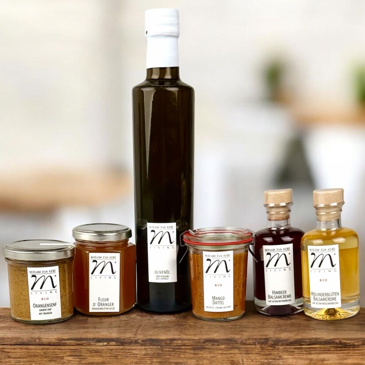 Sechs Feinkostprodukte, bestehend aus drei Gläsern, zwei Flaschen und einer Essigflasche, stehen auf einem Holztisch. Die Produkte tragen weiße Etiketten mit einem stilisierten 'm'. Zu sehen sind u.a. Mangoaufstrich, Blütenhonig, Essig und Olivenöl. Heller, unscharfer Hintergrund.