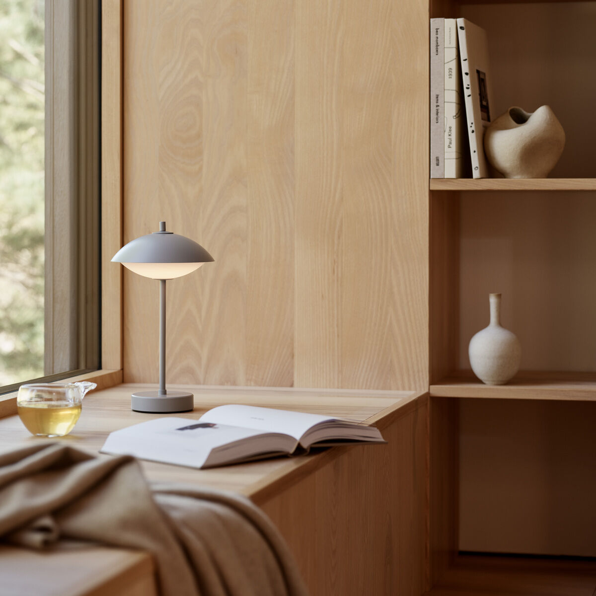 Auf einem fensternahen, hellen Holzsitzplatz steht die graue, tragbare Lampe »Clam™« von Fritz Hansen. Daneben liegen ein aufgeschlagenes Buch, eine Decke und ein Glas Tee. Im Hintergrund befindet sich ein helles Regal mit weißen Vasen und Büchern. Helle, warme Atmosphäre.