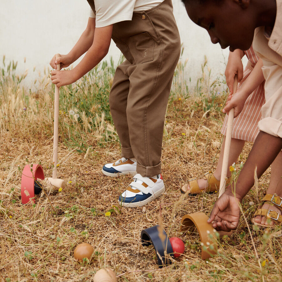 Kinder spielen im Freien Croquet. Ein Kind ist dabei, den Ball mit einem Holzschläger durch einen Torbogen zu schlagen. Verschiedene farbige Bälle und Tore sind auf dem boden verstreut. Das Gras ist trocken und die Kinder sehen konzentriert aus.